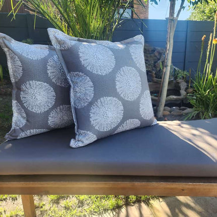 Two gray patterned cushions on a bench outdoors with plants and a blue wall in the background.