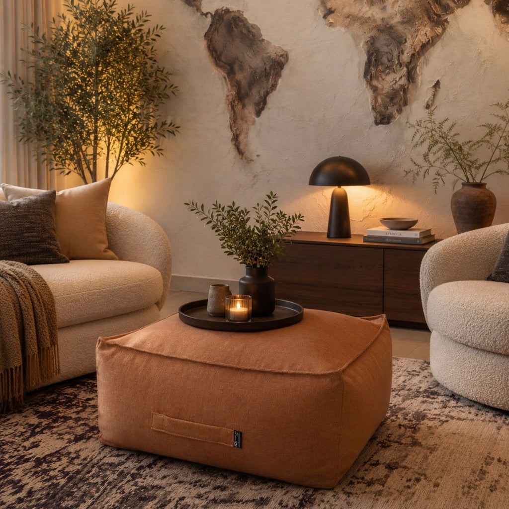 A warm-toned living room scene featuring a large tan fabric pouf ottoman in the foreground, flanked by two cream boucle armchairs. A dark wood sideboard with a black table lamp, books, and potted plants sits against a textured wall with a large world map mural.