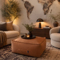 A warm-toned living room scene featuring a large tan fabric pouf ottoman in the foreground, flanked by two cream boucle armchairs. A dark wood sideboard with a black table lamp, books, and potted plants sits against a textured wall with a large world map mural.