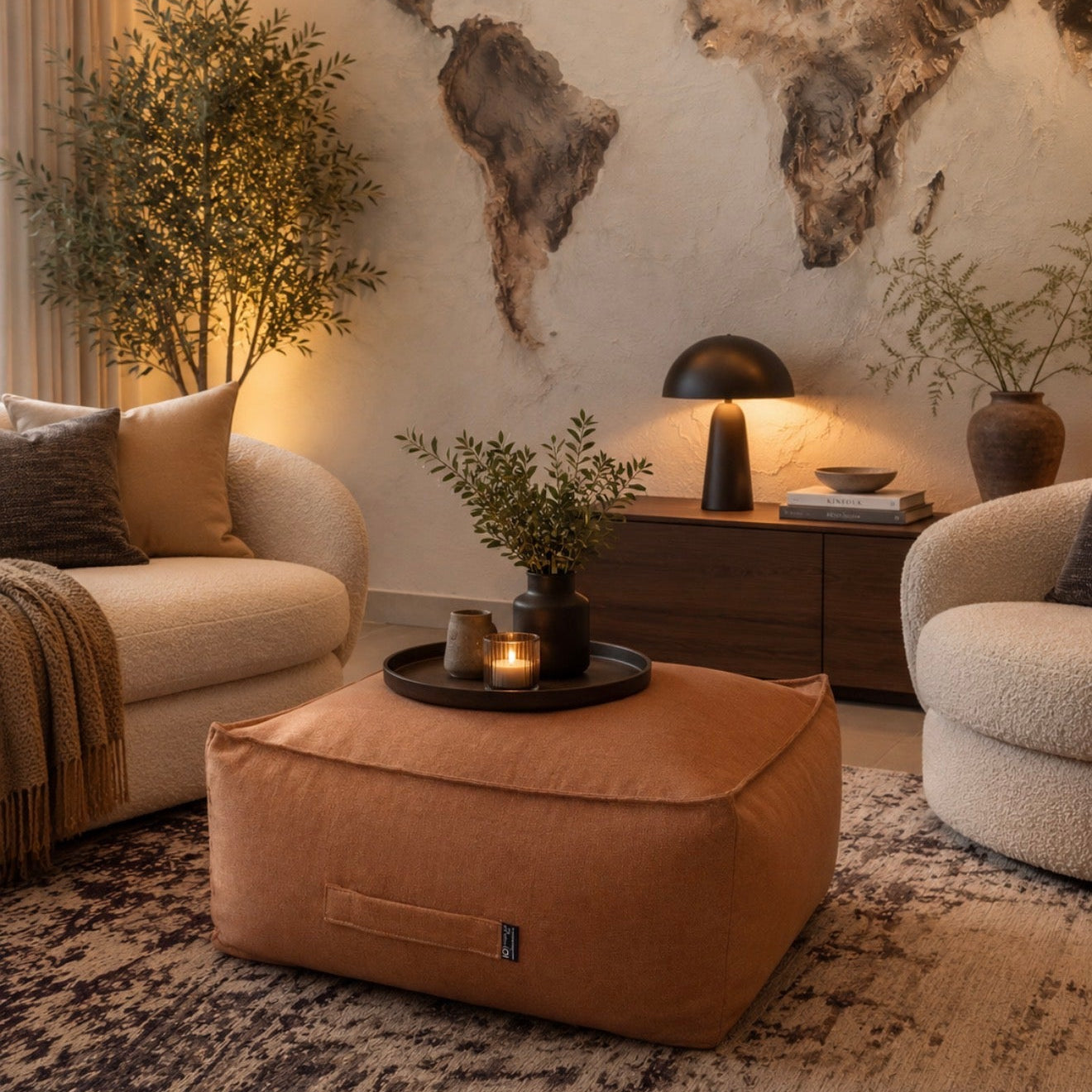 A warm-toned living room scene featuring a large tan fabric pouf ottoman in the foreground, flanked by two cream boucle armchairs. A dark wood sideboard with a black table lamp, books, and potted plants sits against a textured wall with a large world map mural.